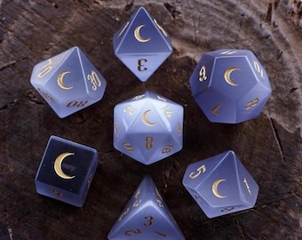 May include: Set of seven purple and white dice with gold crescent moon and number details. The dice are in a variety of shapes, including a d4, d6, d8, d10, d12, and d20.