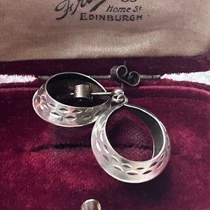 Könnte beinhalten: Silberne Creolen mit einem strukturierten Design. Die Ohrringe befinden sich in einer roten Samtbox mit einem Etikett, auf dem "Watchmaker Jeweller & Optician Francis 53 Home St. EDINBURGH." steht.