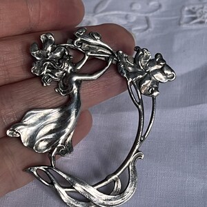 May include: A silver Art Nouveau brooch depicting a woman reaching for flowers. The brooch has a flowing design with detailed floral elements and a curved shape. The piece is a classic example of Art Nouveau jewellery.