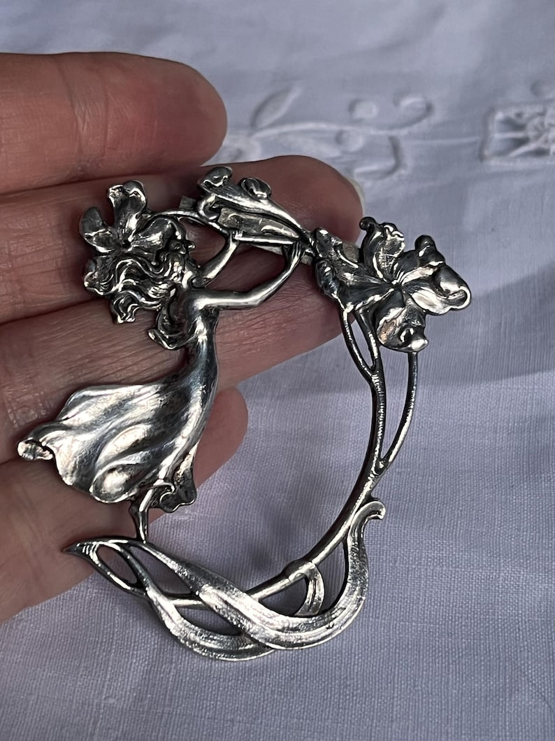 May include: A silver Art Nouveau brooch depicting a woman reaching for flowers. The brooch has a flowing design with detailed floral elements and a curved shape. The piece is a classic example of Art Nouveau jewellery.