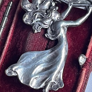 May include: A silver Art Nouveau brooch featuring a woman with flowing hair and a floral motif. The brooch is set against a deep red velvet background, highlighting the intricate details and classic design.