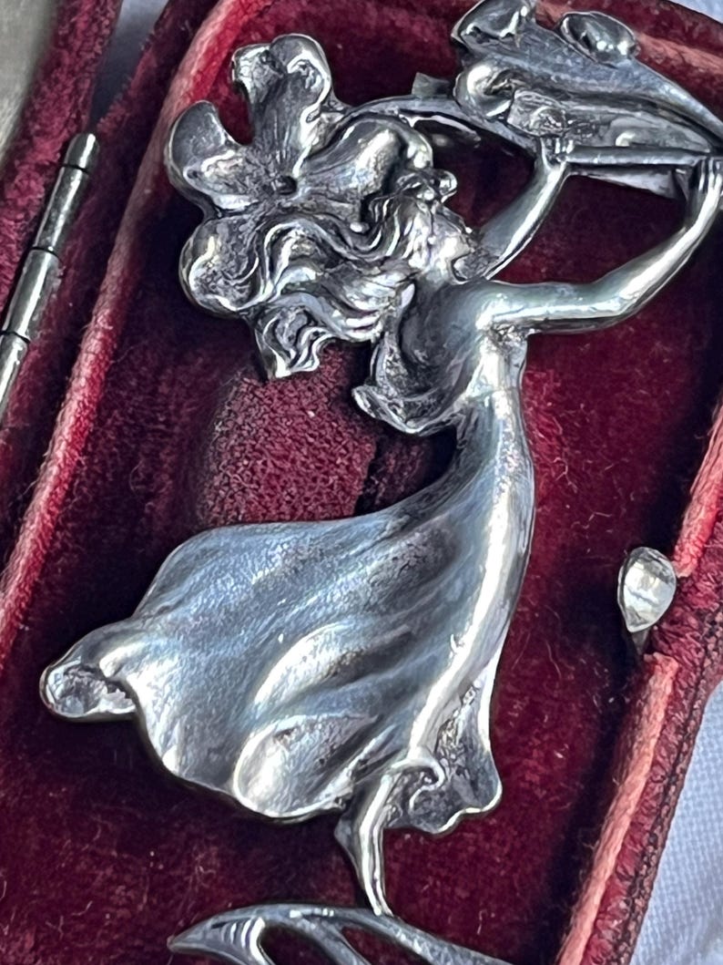May include: A silver Art Nouveau brooch featuring a woman with flowing hair and a floral motif. The brooch is set against a deep red velvet background, highlighting the intricate details and classic design.