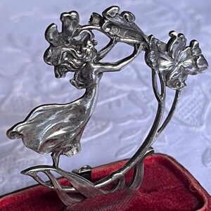 May include: A silver Art Nouveau brooch depicting a woman reaching for flowers. The brooch has detailed features and a flowing design. It is set against a red velvet background.