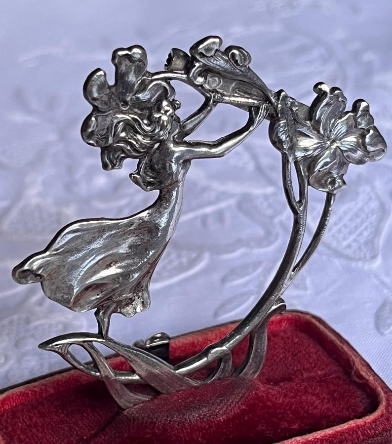 May include: A silver Art Nouveau brooch depicting a woman reaching for flowers. The brooch has detailed features and a flowing design. It is set against a red velvet background.