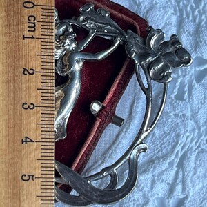 May include: A silver Art Nouveau brooch depicting a woman and flowers. The brooch is approximately 5 cm long and set against a red velvet background. The design features floral and flowing lines, typical of the style.