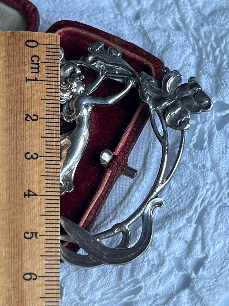 May include: A silver Art Nouveau brooch depicting a woman and flowers. The brooch is approximately 5 cm long and set against a red velvet background. The design features floral and flowing lines, typical of the style.