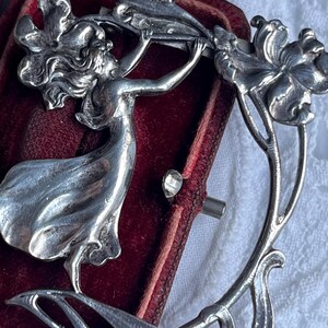 May include: A silver Art Nouveau brooch depicting a woman reaching for flowers. The design features flowing lines and floral motifs. The brooch is set against a red velvet background.