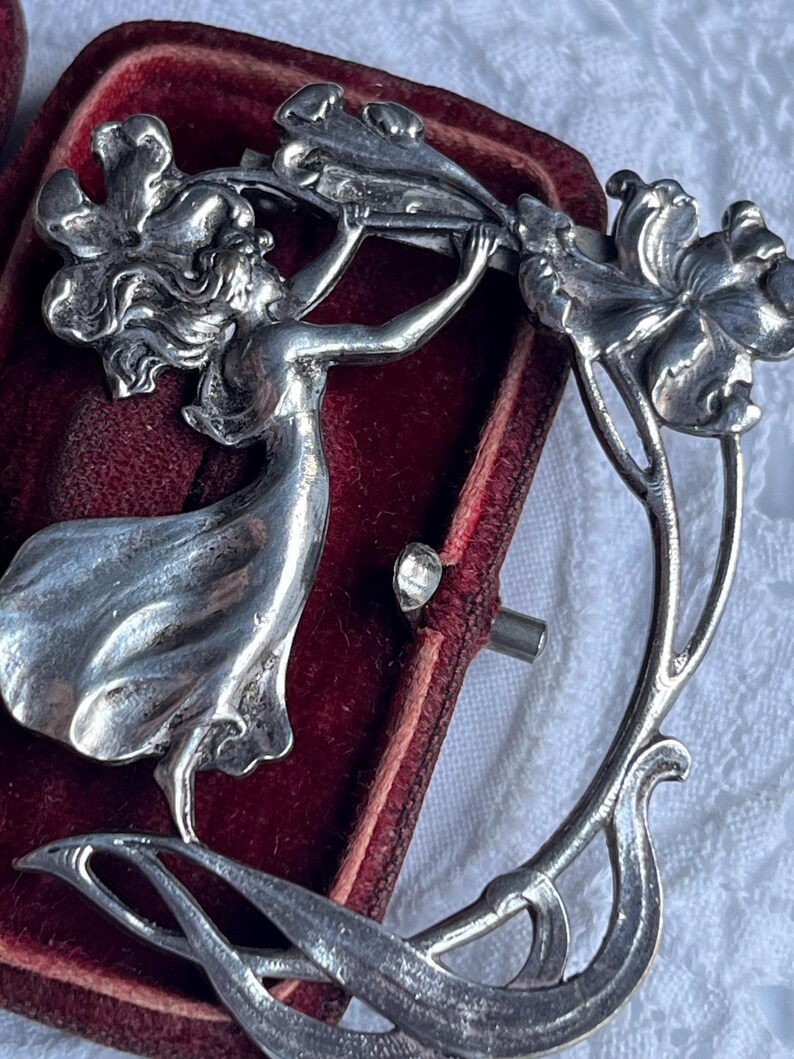 May include: A silver Art Nouveau brooch depicting a woman reaching for flowers. The design features flowing lines and floral motifs. The brooch is set against a red velvet background.