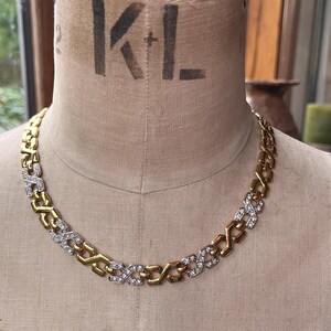 Könnte beinhalten: Eine goldfarbene Kette mit abwechselnd goldenen und silberfarbenen Gliedern. Die silbernen Glieder sind mit kleinen, klaren Kristallen verziert. Die Halskette wird auf einer Schaufensterpuppe präsentiert.