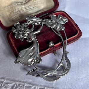 May include: A silver Art Nouveau brooch depicting a woman and flowers. The brooch is displayed in a red velvet box. The design features flowing lines and floral motifs, reflecting a vintage aesthetic. The brooch is a classic piece of jewellery.