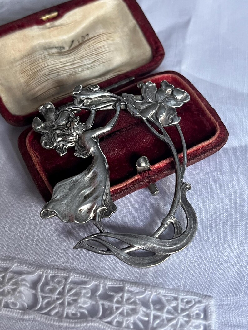 May include: A silver Art Nouveau brooch depicting a woman and flowers. The brooch is displayed in a red velvet box. The design features flowing lines and floral motifs, reflecting a vintage aesthetic. The brooch is a classic piece of jewellery.