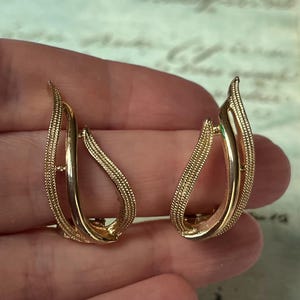 Könnte beinhalten: Ein Paar goldfarbene Clip-Ohrringe. Jeder Ohrring hat ein gebogenes, offenes Design mit einem strukturierten, perlenbesetzten Außenrand und einer glatten, durchgehenden Innenkurve. Die Ohrringe haben eine blattähnliche Form.