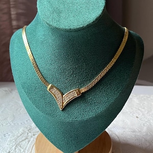 Puede incluir: Un collar de cadena dorada con un colgante en forma de V engastado con diamantes de imitación transparentes. El collar se muestra en un busto de terciopelo verde.