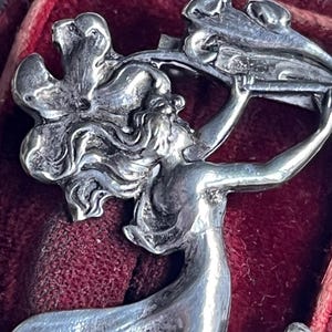 May include: A close-up of a silver brooch. The brooch features a woman holding a floral design. The woman has long flowing hair and is wearing a flowing gown. The brooch is set against a red velvet background.
