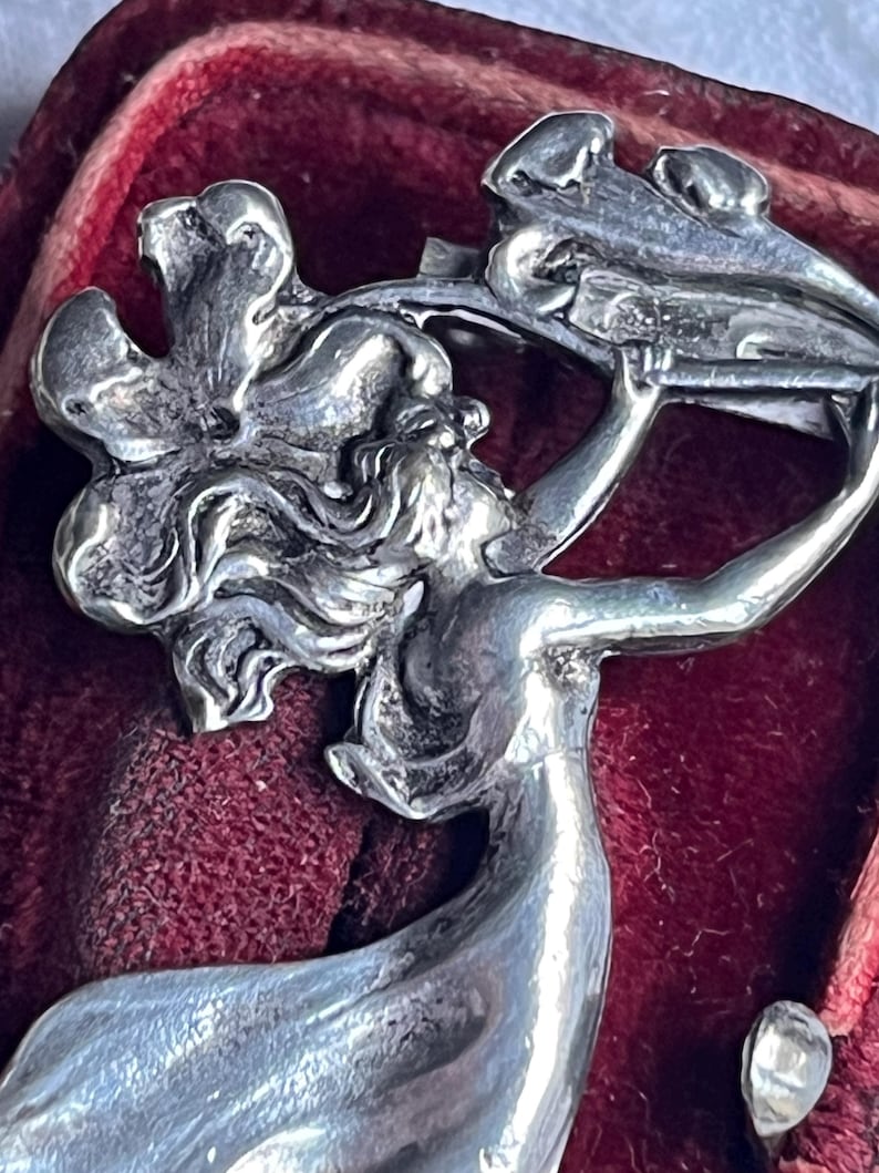 May include: A close-up of a silver brooch. The brooch features a woman holding a floral design. The woman has long flowing hair and is wearing a flowing gown. The brooch is set against a red velvet background.
