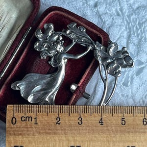 May include: A silver Art Nouveau brooch depicting a woman with flowing hair and dress, holding a floral design. The brooch is approximately 5 cm long and is displayed in a red velvet box.