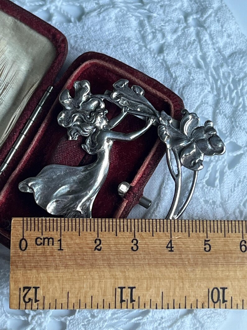 May include: A silver Art Nouveau brooch depicting a woman with flowing hair and dress, holding a floral design. The brooch is approximately 5 cm long and is displayed in a red velvet box.