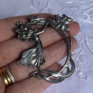 May include: A silver Art Nouveau brooch depicting a woman with flowers. The brooch has a flowing design with floral details. The metal has a slightly aged appearance. The brooch is a decorative accessory.