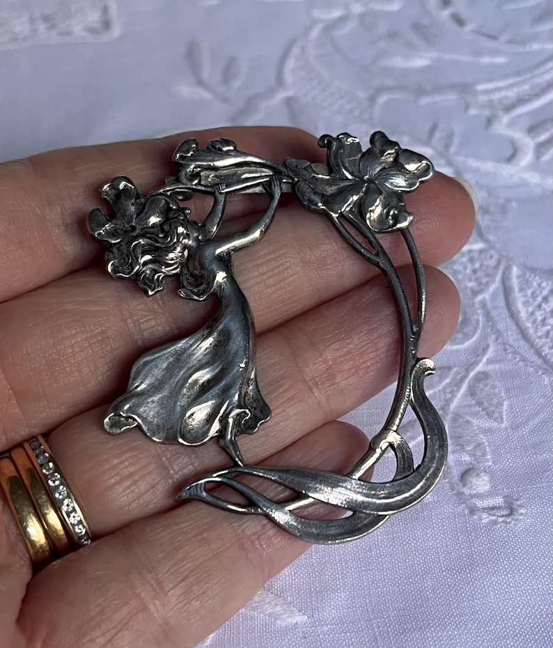May include: A silver Art Nouveau brooch depicting a woman with flowers. The brooch has a flowing design with floral details. The metal has a slightly aged appearance. The brooch is a decorative accessory.