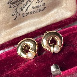 Könnte beinhalten: Ein Paar goldfarbene Ohrringe mit Perlenakzent. Die Ohrringe befinden sich in einer roten Samt-Schmuckschatulle mit dem Text "J. Frame, Jeweller & Optician, Home St. Edinburgh".