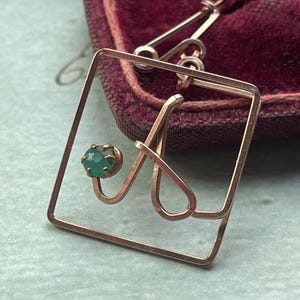 Könnte beinhalten: Ein goldfarbener Anhänger mit einem grünen Edelstein, der in einen quadratischen Rahmen gefasst ist. Der Anhänger ist aus Draht gefertigt und hat ein filigranes, kompliziertes Design.