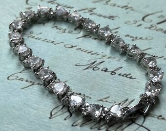 May include: A silver bracelet featuring heart-shaped clear gemstones. The bracelet is made of silver-toned metal and has a clasp closure. The gemstones are evenly spaced along the bracelet's length, creating a sparkling effect.