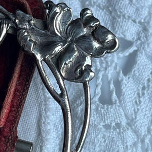 May include: Close-up of a silver floral hair accessory. The detailed design features a flower with multiple petals and a stem. The accessory is likely vintage or antique, with a classic, elegant style.