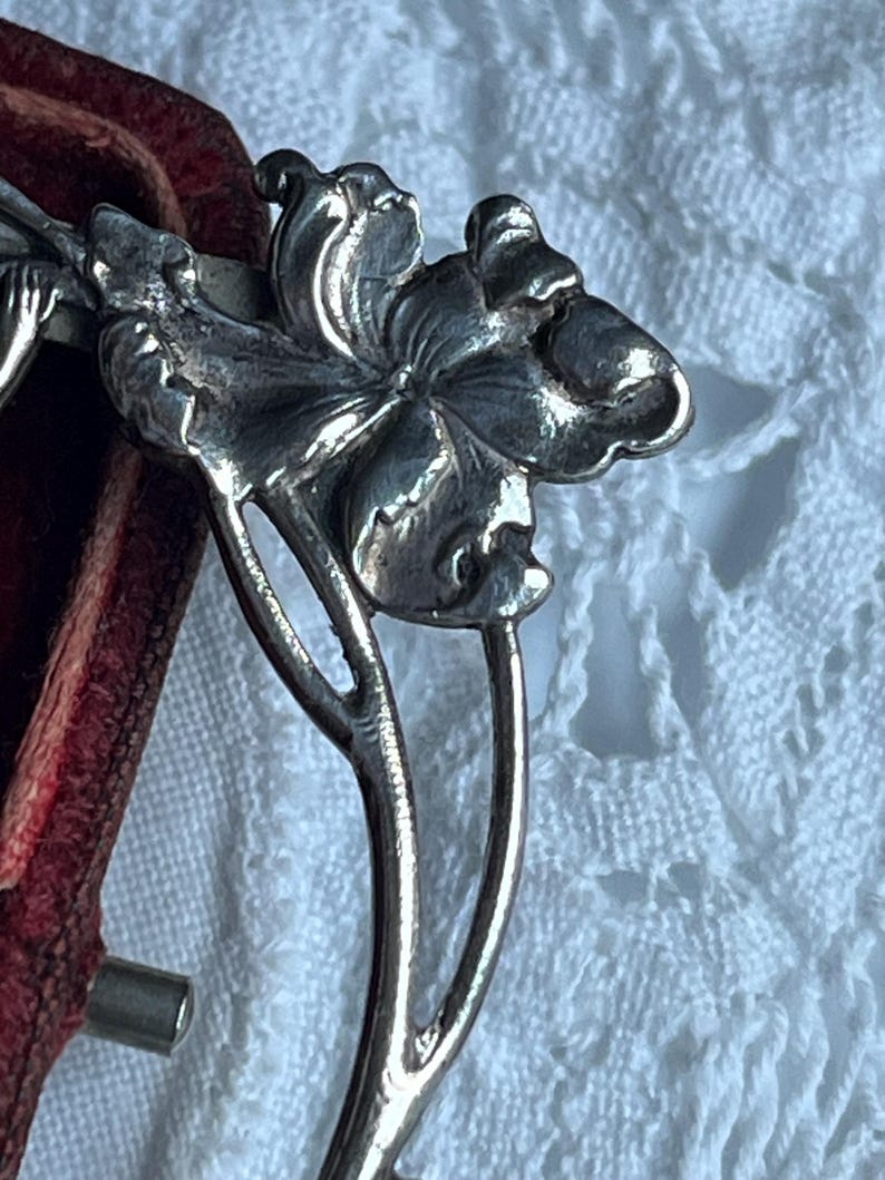 May include: Close-up of a silver floral hair accessory. The detailed design features a flower with multiple petals and a stem. The accessory is likely vintage or antique, with a classic, elegant style.