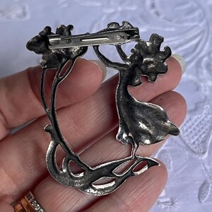 May include: A silver brooch depicting a woman reaching for a floral element. The brooch has a curved design with detailed leaves and flowers. The pin closure is visible on the back, crafted in a silver tone.