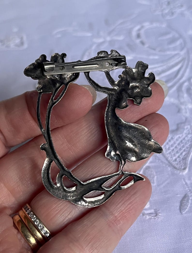 May include: A silver brooch depicting a woman reaching for a floral element. The brooch has a curved design with detailed leaves and flowers. The pin closure is visible on the back, crafted in a silver tone.