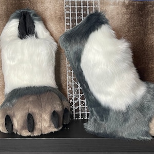 May include: Pair of furry, plush paw-shaped costume feet. The paws are primarily white with gray and brown accents. Each paw has four black claws. The feet are designed to be worn as part of a costume.