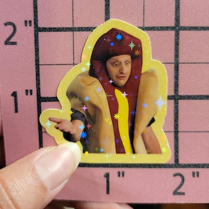 May include: A holographic sticker featuring a person dressed as a hot dog. The person is wearing a red bun hat with star accents and a yellow hot dog costume. The sticker is held by a hand.