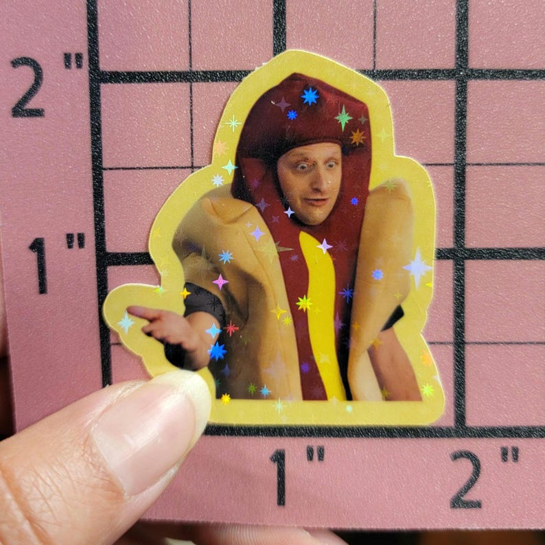 May include: A holographic sticker featuring a person dressed as a hot dog. The person is wearing a red bun hat with star accents and a yellow hot dog costume. The sticker is held by a hand.