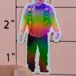 May include: A holographic sticker of a man in a button-down shirt, vest, and pants. The sticker is approximately 2 inches tall. The image has a rainbow gradient effect, with the man's face and hands in a lighter color.