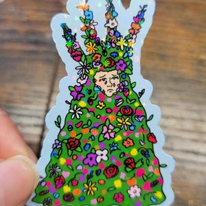 May include: A holographic sticker featuring a person covered in green foliage and colorful flowers. The person has a sad expression. The sticker has a light blue border and is held by a hand.