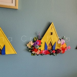 May include: Three yellow triangular wooden structures with blue accents and a small window. The middle structure is decorated with a garland of colorful artificial flowers.