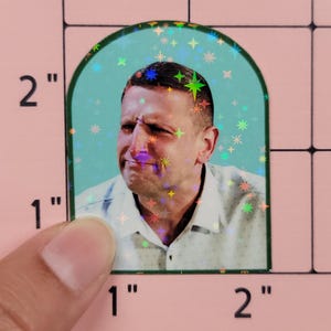 May include: A holographic sticker featuring a man's face within a teal arch, surrounded by star-shaped sparkles. The man is wearing a white collared shirt. The sticker is approximately 2 inches tall and 1 inch wide.