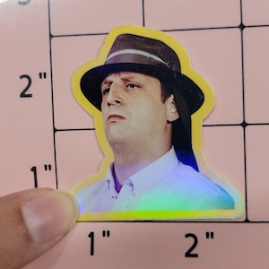 May include: A sticker featuring a man wearing a black fedora hat with a brown brim. The sticker has a yellow border and a holographic effect. The man is wearing a white collared shirt. The sticker is approximately 2 inches tall.