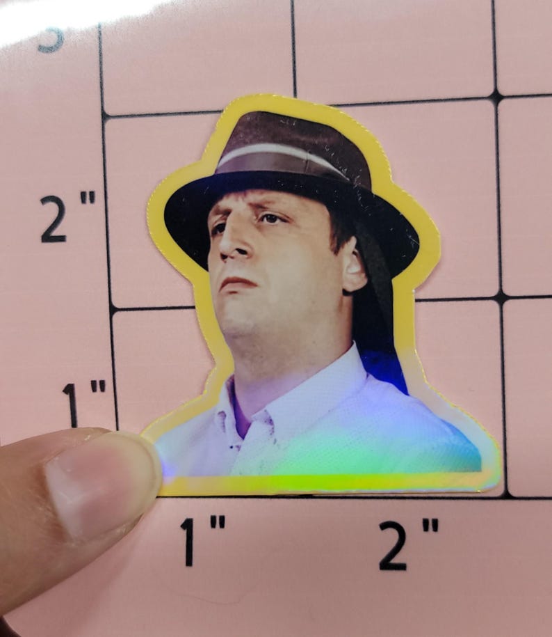 May include: A sticker featuring a man wearing a black fedora hat with a brown brim. The sticker has a yellow border and a holographic effect. The man is wearing a white collared shirt. The sticker is approximately 2 inches tall.