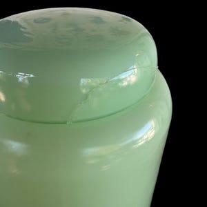 May include: A pale green glass jar with a lid, showing a crack. The jar is set against a black background. The jar is likely vintage.