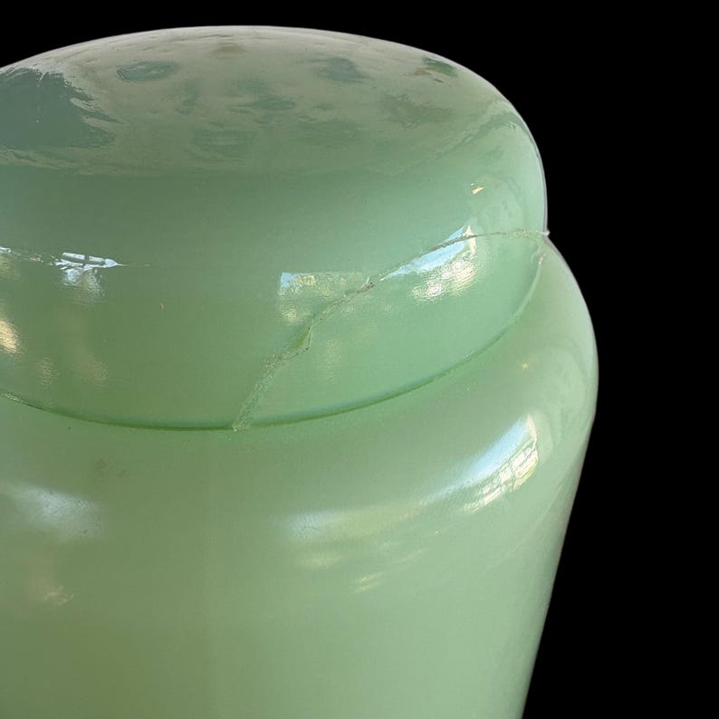 May include: A pale green glass jar with a lid, showing a crack. The jar is set against a black background. The jar is likely vintage.