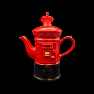 May include: A red ceramic teapot shaped like a British postbox, with a crown on top. The teapot has a black base with a decorative emblem. The spout and handle are curved, and the design includes a small door and a yellow and black slot.