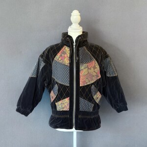 May include: Black velvet jacket with floral and geometric patchwork. The jacket features a high collar, a full-length zipper, and a cinched waist. The patchwork includes shades of pink, green, and blue.