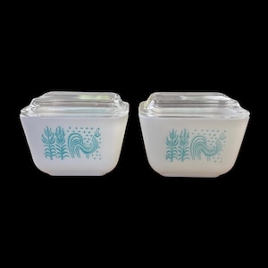 Peut inclure: Deux plats de conservation carrés en verre opalin blanc avec des couvercles en verre transparent. Chaque plat présente un motif graphique bleu sarcelle représentant un coq à côté de tiges de maïs, agrémenté de petites croix au-dessus.
