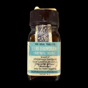 May include: A vintage amber glass medicine bottle with a dark brown screw-top lid. The label reads 100 ORAL TABLETS LEVO-DROMORAN TARTRATE ROCHE, Brand of LEVORPHAN TARTRATE, Warning may be habit forming, and HOFFMANN-LA ROCHE INC. NUTLEY, N. J.