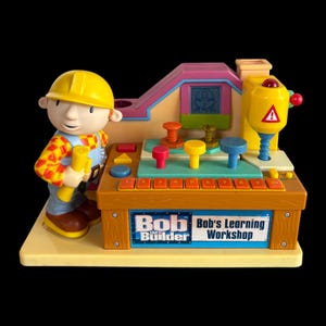 Op de afbeelding: Een Bob de Bouwer leerwerkplaats speelgoed van kunststof met een figuur in een gele helm die een gele bouwtekening vasthoudt. De console bevat een boor, gekleurde pinnen, een rij genummerde knoppen van 1 tot 10 en een scherm met een icoon.