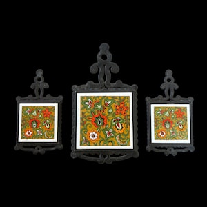 May include: A set of three black cast iron trivets with ornate scrollwork handles and square ceramic inserts. Each insert features a vibrant floral pattern with orange, red, and green flowers set against a mustard yellow background.
