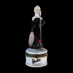 May include: A porcelain Barbie figurine in a black gown with a microphone, standing on a white and gold box. The box is labeled "Barbie SOLD IN THE SPOTLIGHT". The figurine has blonde hair and red lipstick.