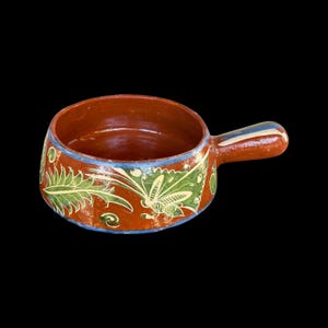 May include: A small, round, terracotta-colored ceramic bowl with a handle. The bowl is decorated with green and white floral and insect designs, outlined in blue. The handle has blue and white stripes.