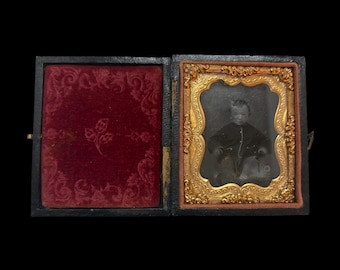 May include: A black and white daguerreotype of a young child in a dark jacket, sitting in a chair. The image is framed in a gold-colored frame with ornate details. The frame is in a black wooden case with a red velvet lining.